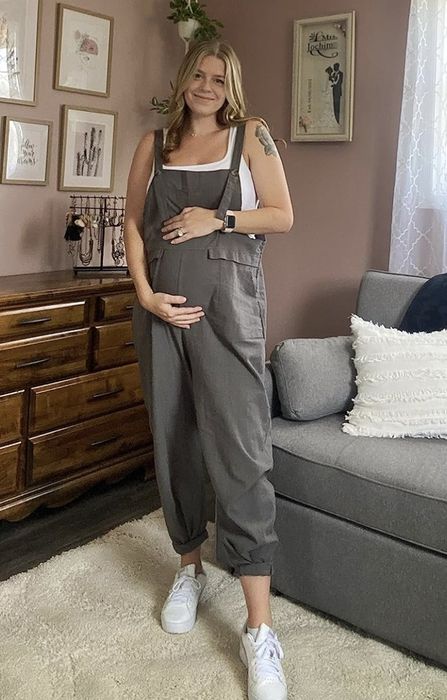 A smiling pregnant woman stands in her living room, wearing overalls and sneakers, one hand resting on her belly. Art and decor pieces adorn the background.