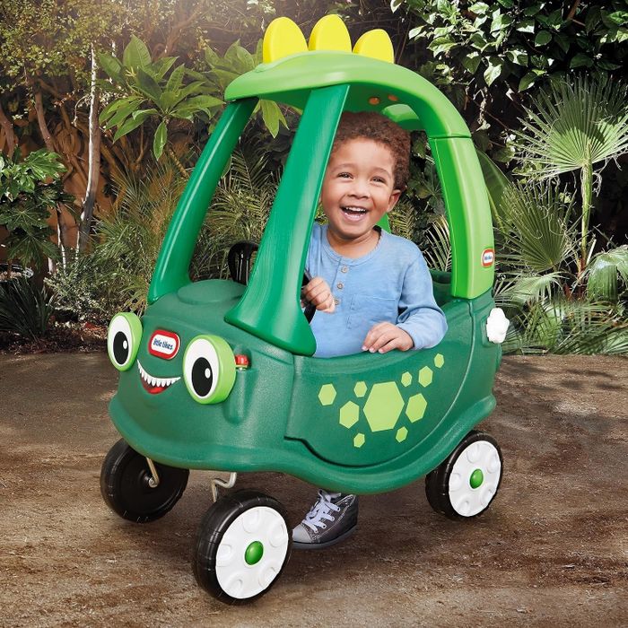 A child sitting happily in a green, dinosaur-themed toy car, complete with yellow spikes and hexagonal patterns, set against a forest-like backdrop