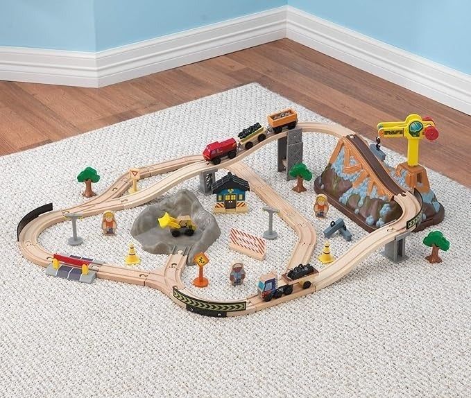 A wooden toy train set on a white carpet, featuring tracks, trees, a mountain tunnel, a station, and various accessories such as signs and figures