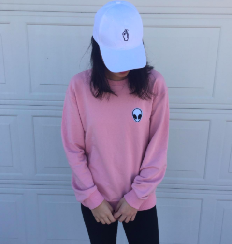 The reviewer is wearing the same style of sweatshirt in pink, along with a white baseball cap and leggings.