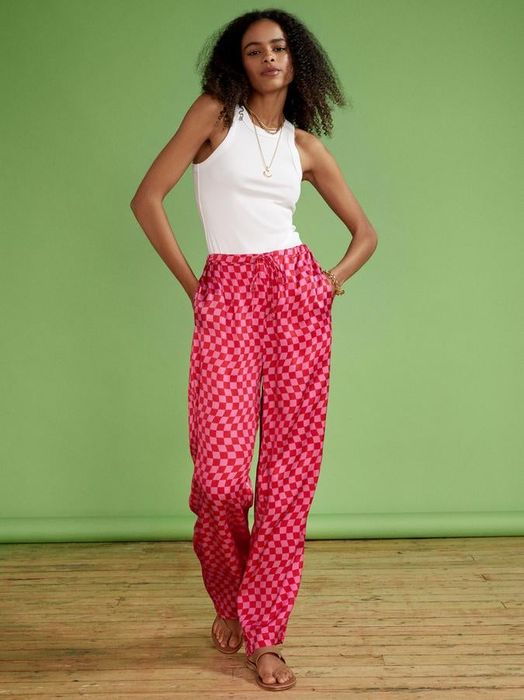 Model showing off bright pink checkerboard trousers, styled with tan sandals and a white tank top for a relaxed, summery look.