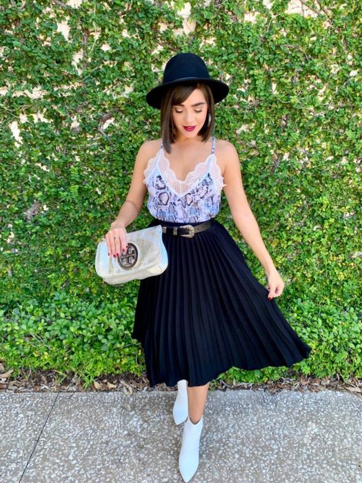 The reviewer is dressed in a black pleated skirt paired with a cami, white boots, and a fedora.