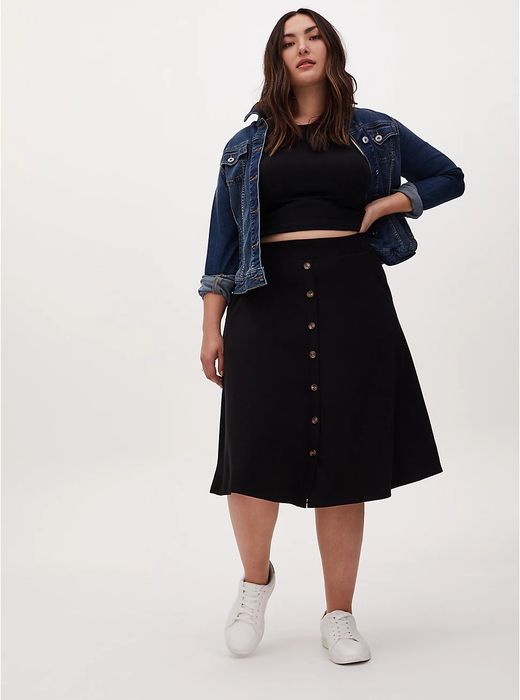 A plus-size model sporting a black knee-length skirt, featuring brown buttons running down the center.