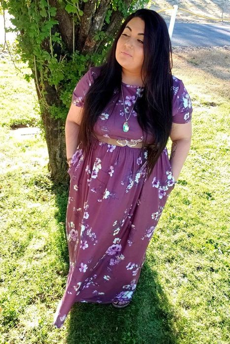 The reviewer is wearing a purple floral print dress, accessorized with a belt around the waist.