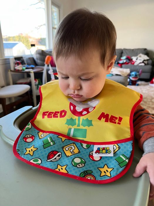 A baby wearing a bib with a Mario theme that reads 'Feed me'