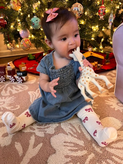 A baby sitting on a rug, chewing on a toy giraffe, in front of a beautifully decorated Christmas tree. The baby is dressed in a cute dress and patterned tights