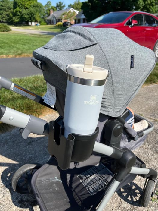 A stroller with a gray canopy and a cup holder holding a Stanley travel tumbler, positioned along an outdoor path