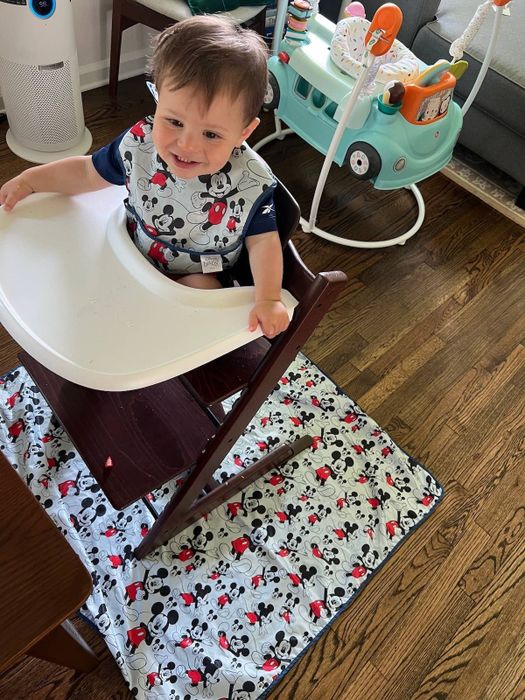A baby sitting in a high chair with a Mickey Mouse bib and matching floor mat. A great fit for Disney-loving families