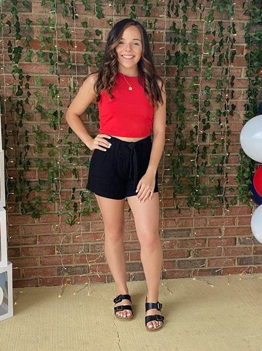 A reviewer is seen wearing a red top with shorts and sandals.