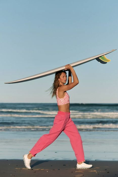 Model strolling along the beach, carrying a surfboard above her head, wearing pink pants