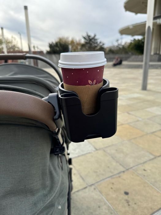 A stroller with a cup holder on the handle, holding a takeaway coffee cup with a polka-dot sleeve, set in an outdoor environment