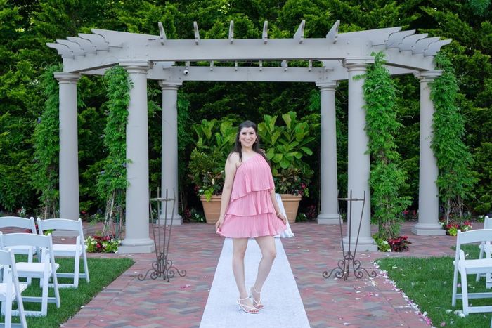 A reviewer stands beneath a pergola on a white aisle at an outdoor venue, wearing a short, layered, sleeveless dress and white shoes