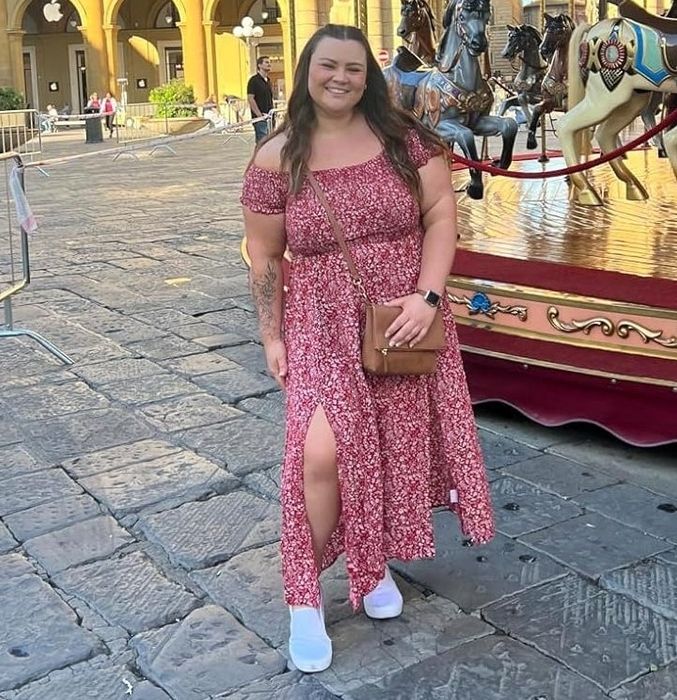 A reviewer beams in front of a beautifully ornate European carousel, dressed in a red patterned off-shoulder dress, white shoes, and holding a brown bag