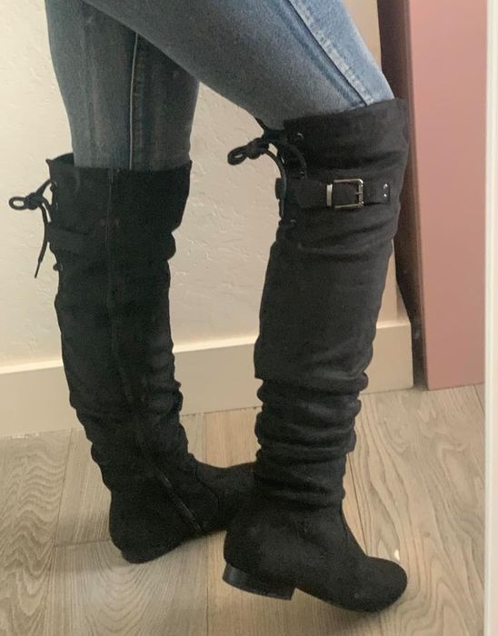 A close-up of a person wearing black suede knee-high boots with buckle accents, paired with jeans, standing indoors near a wall.