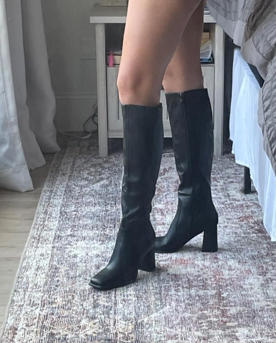 A person stands indoors, showcasing stylish knee-high black leather boots, positioned next to a rug and a bed.