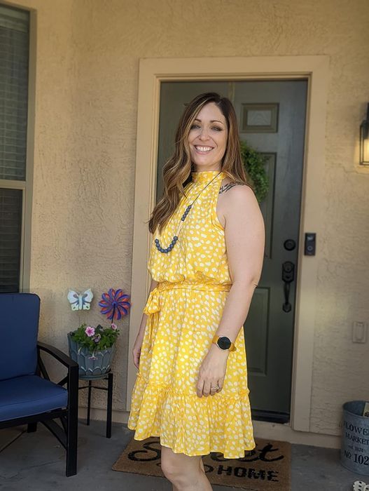 Woman in a yellow patterned sleeveless dress and necklace, smiling on a porch of a house