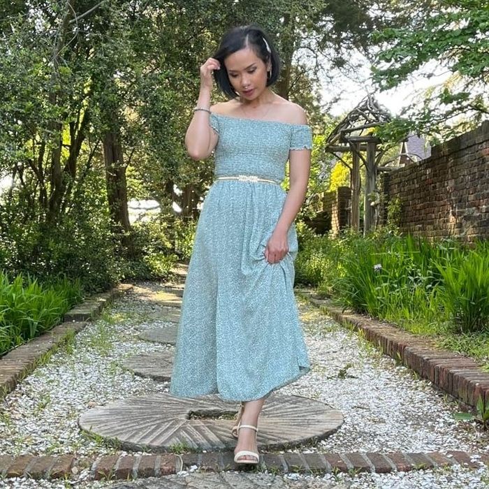 A reviewer stands on a stone path in a garden, wearing a light green floral midi dress with an off-shoulder design