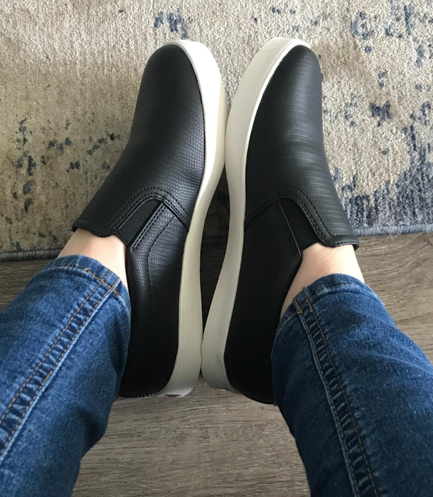 A person stands on a textured surface, wearing relaxed-fit blue jeans and black slip-on sneakers with white soles.