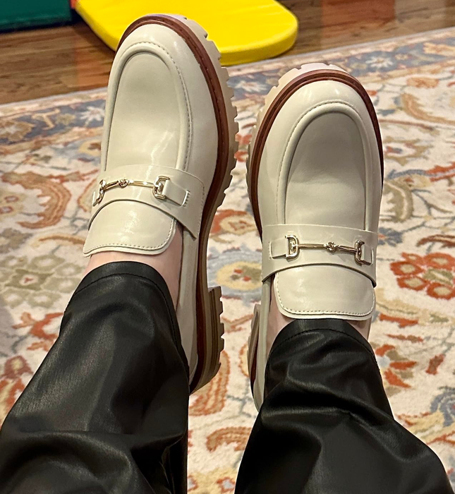 A person seated indoors, wearing glossy black pants and cream-colored platform loafers with gold buckle embellishments.