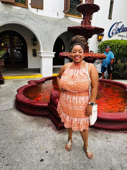 Woman in a patterned dress and sandals, smiling by a fountain while holding a white purse