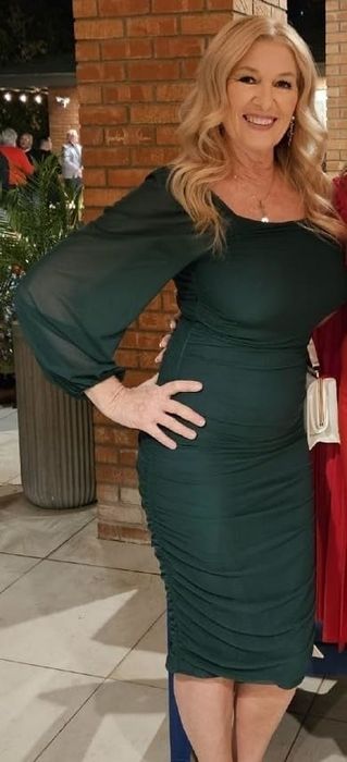 A smiling reviewer poses in front of a brick wall and potted plants, wearing a fitted long-sleeve dress with ruched detailing, one hand on her hip