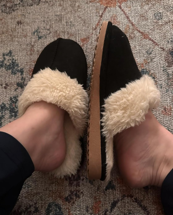 Two feet are shown in fluffy beige slip-on slippers with black uppers, resting on a patterned carpet.