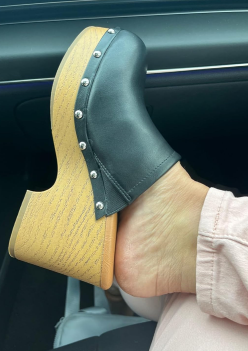 A person’s foot showcasing a wooden platform clog with black leather and silver studs, paired with light-colored trousers.