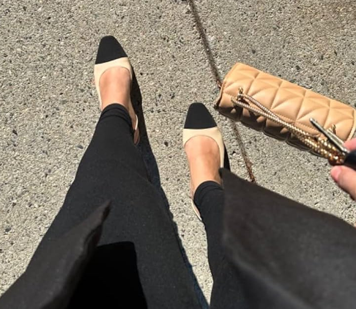 A person wearing black flats, holding a quilted clutch purse, and standing on the pavement.