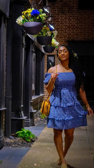 Woman wearing a blue ruffled dress, carrying a shoulder bag, strolling down a city sidewalk at night