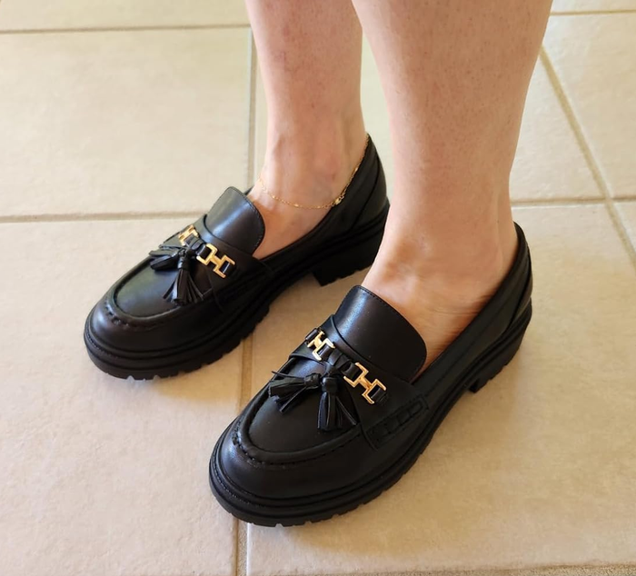 A close-up of someone's feet adorned in black loafers with tassel accents and gold detailing, standing on a patterned tile floor.
