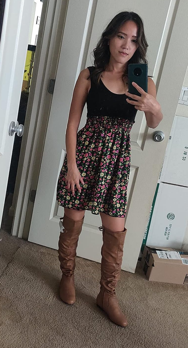 A reviewer poses in a mirror selfie, wearing a black floral dress and brown knee-high boots in a cozy room.