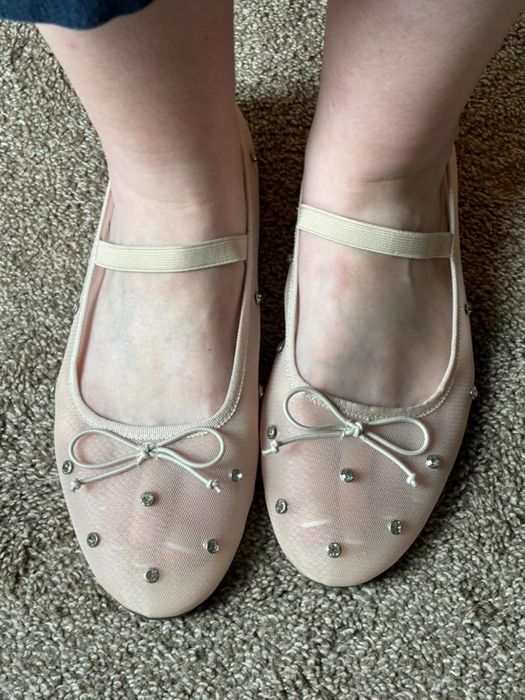 A close-up of feet wearing beige Mary Jane flats, complete with decorative bows and small metallic studs, displayed on a textured surface.
