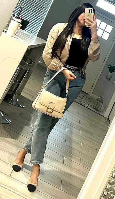 A reviewer in a stylish casual outfit, featuring jeans, a black top, a beige jacket, and a beige handbag, taking a mirror selfie in a contemporary home setting.