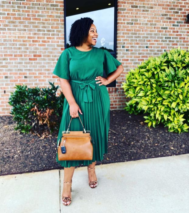 A reviewer wearing a pleated dress, smiling and holding a brown handbag.