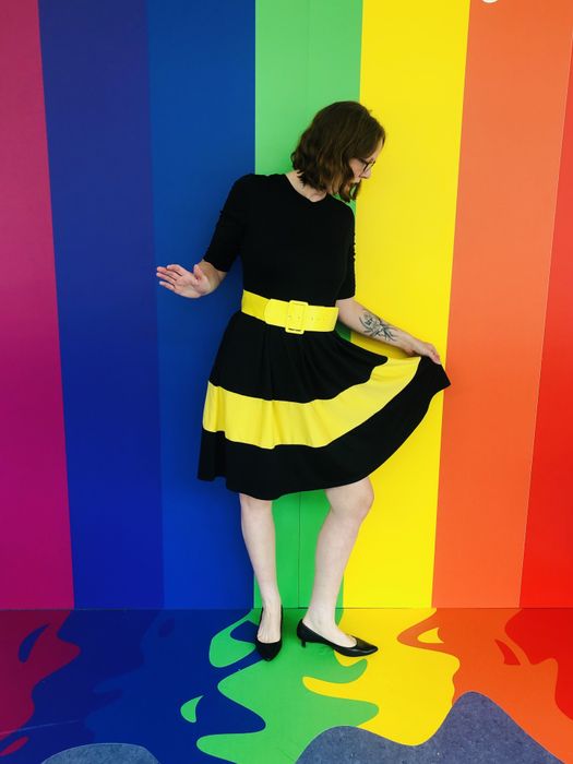 A reviewer wearing a black dress with a yellow stripe, posing in front of a vibrant rainbow wall.