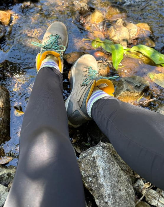 A person’s legs wearing hiking boots and dark leggings, standing on rocks next to a stream.