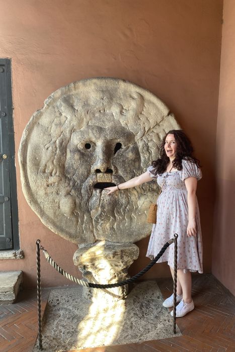 A writer wearing the same dress, with their hand placed in the mouth of a statue.