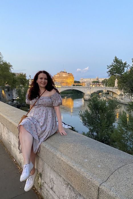 A writer in a midi-length dress with off-the-shoulder puff sleeves, casually seated on the wall of a bridge.