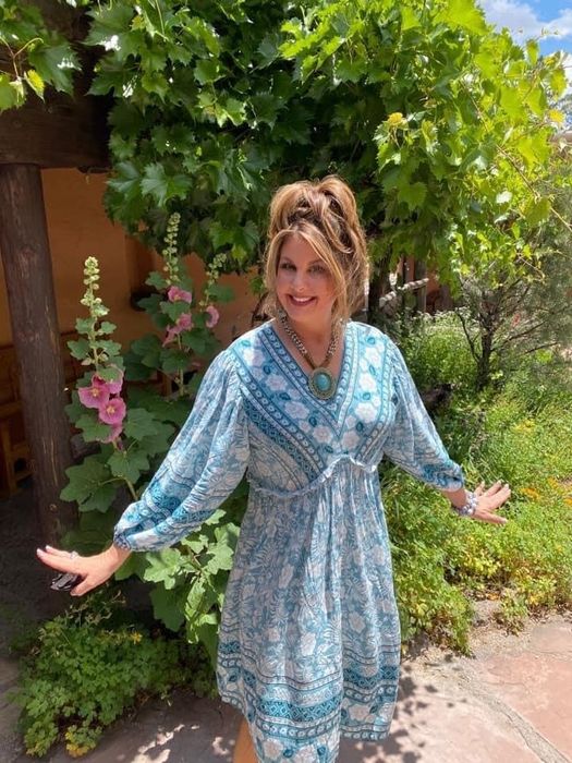 Woman in a patterned dress, arms outstretched, standing under a pergola in a sunlit garden