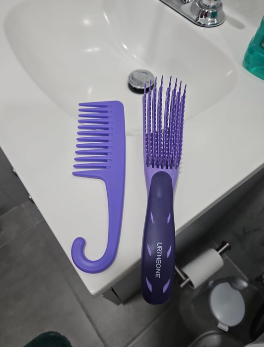 Two purple hair tools resting on a bathroom sink: a wide-tooth comb with a hook handle and a detangling brush labeled 'URTHEONE.'