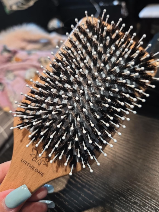 A wooden paddle hairbrush with black bristles and white ball tips, held in a hand with painted nails.