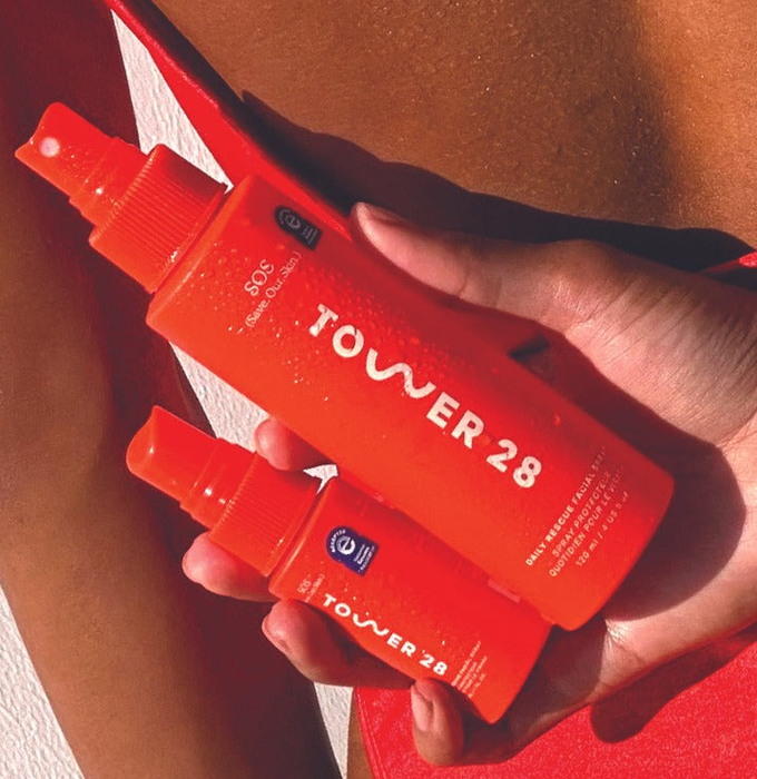 A model holding both the mini and full-sized bottles of Tower 28 SOS Daily Rescue Facial Spray in one hand.