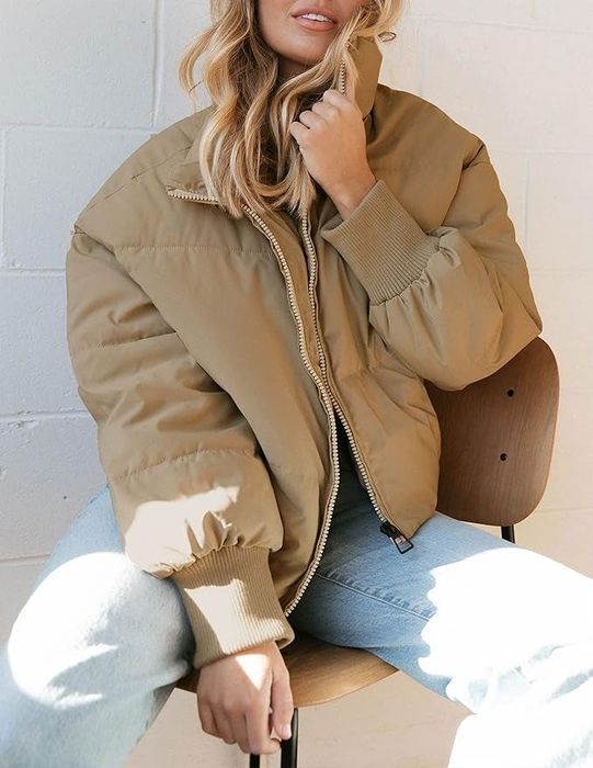 Person rocking a stylish oversized puffer jacket, partially zipped, paired with light blue jeans, seated on a wooden chair indoors