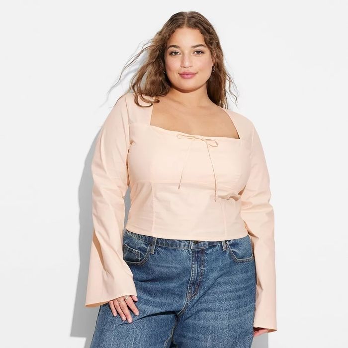 Woman wearing a long-sleeved, square-neck top with a tie detail and relaxed-fit jeans, posing confidently. Fashion photography.