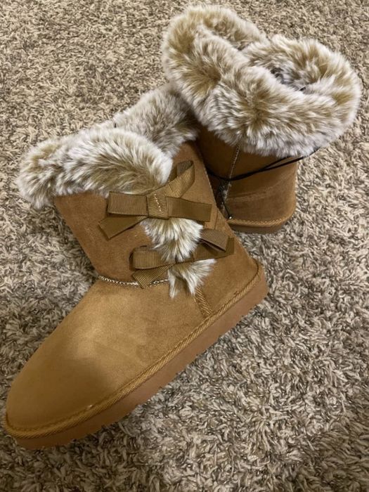 Faux-fur-lined suede boots with ribbon bows, elegantly displayed on a textured carpet, highlighted in a shopping article for their stylish appeal.