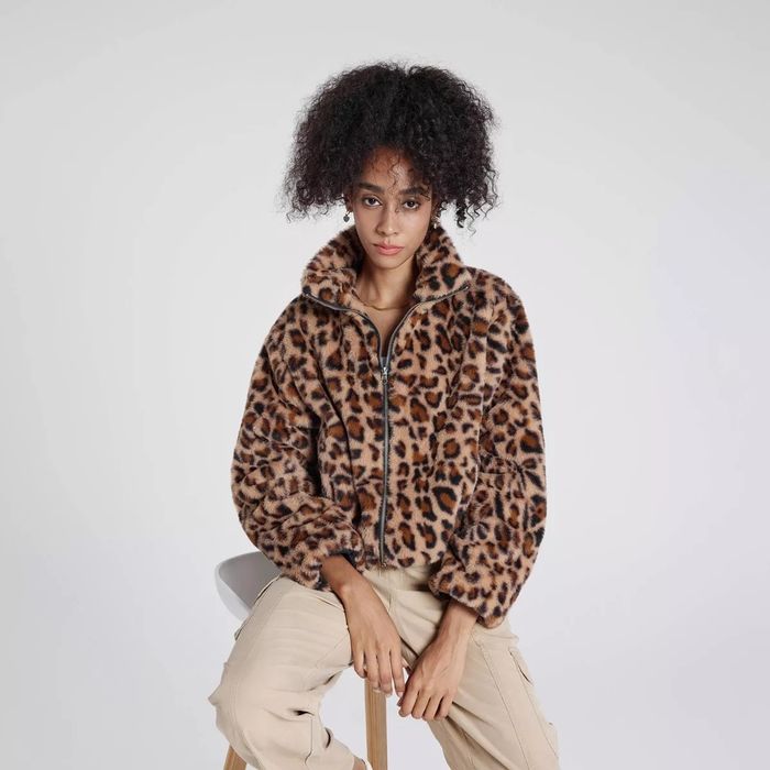 A person sits comfortably wearing a leopard print jacket paired with beige cargo pants, gazing at the camera. The stylish outfit is featured in a shopping article.