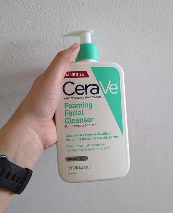A reviewer holds a 16 fl oz pump bottle of CeraVe Foaming Facial Cleanser for normal to oily skin, with text highlighting its benefits and ingredients.