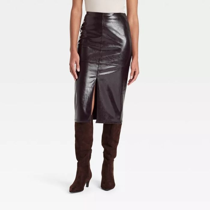 Woman modeling a glossy black pencil skirt with a front slit and knee-high brown boots, set against a simple backdrop.