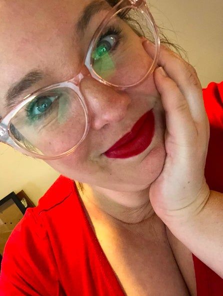 A reviewer, wearing bold red lipstick and glasses, gently touches her face while gazing directly at the camera.