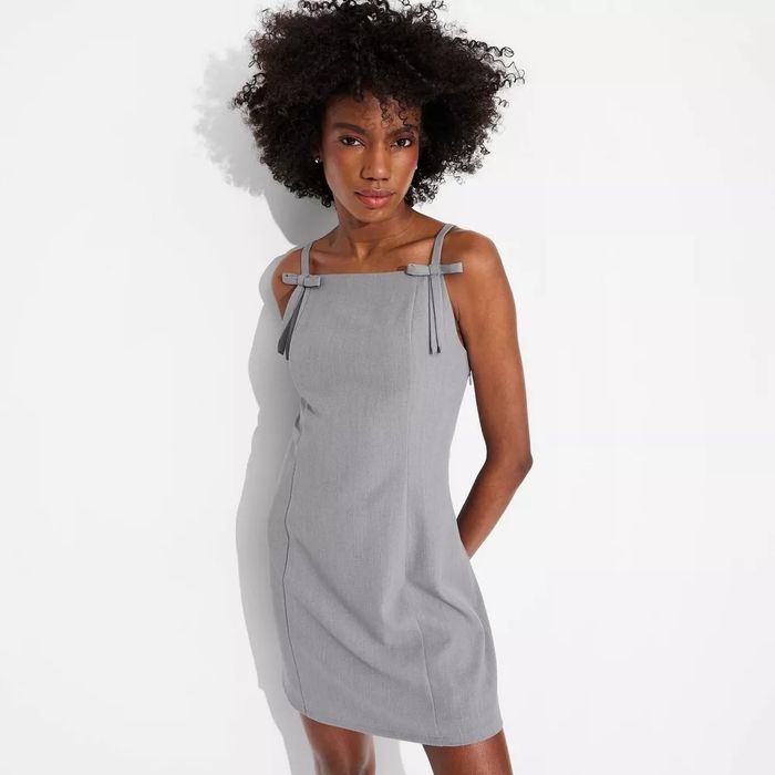 Person wearing a sleeveless, fitted gray dress with bow details on the straps, striking a pose against a simple background.
